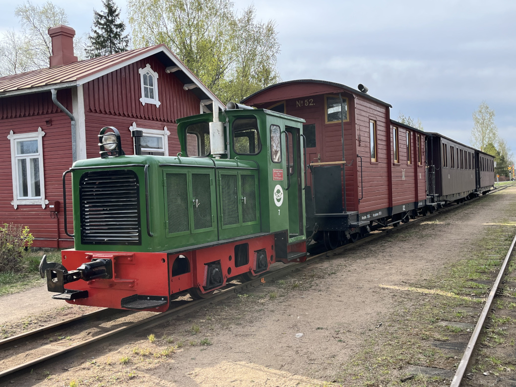 Pienen vihreän dieselveturin vetämä komivaunuinen matkustajajuna punaisen puisen Minkiön asemarakennuksen edessä.